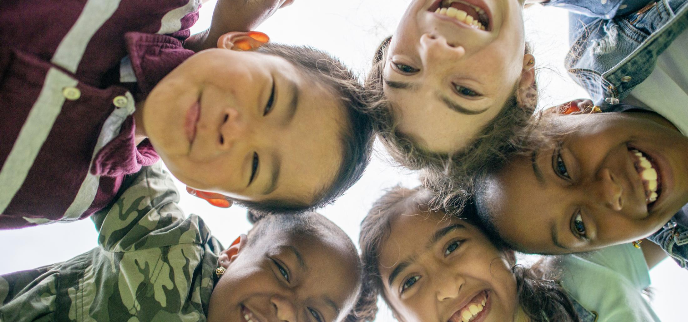 Five children with their arms around each other put their heads together and smile down at the camera.