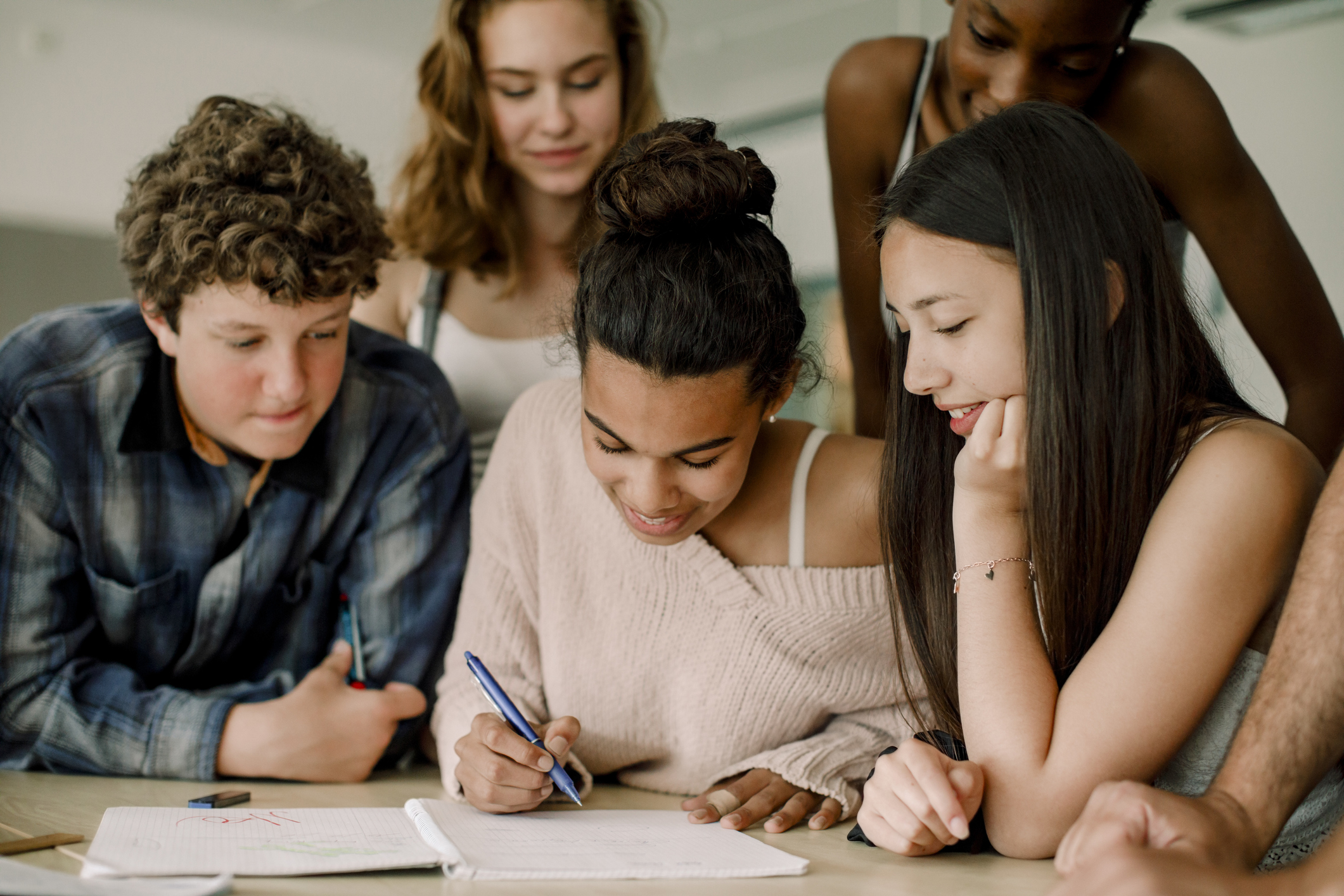 Smiling teenage student writing in notebook while sitting amidst friends in classroom.