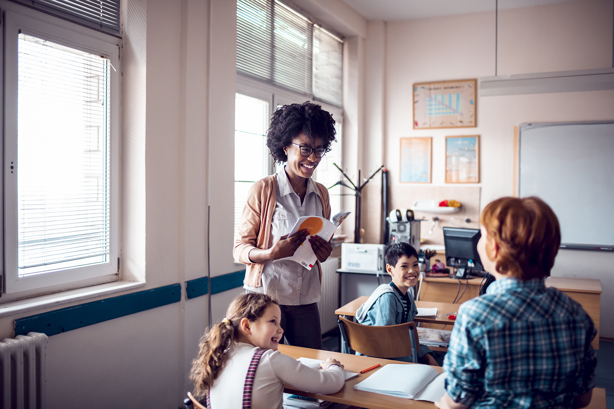 A teacher smiles at a student who is standing in the classroom. Two other students are looking and smiling at the student, as well.