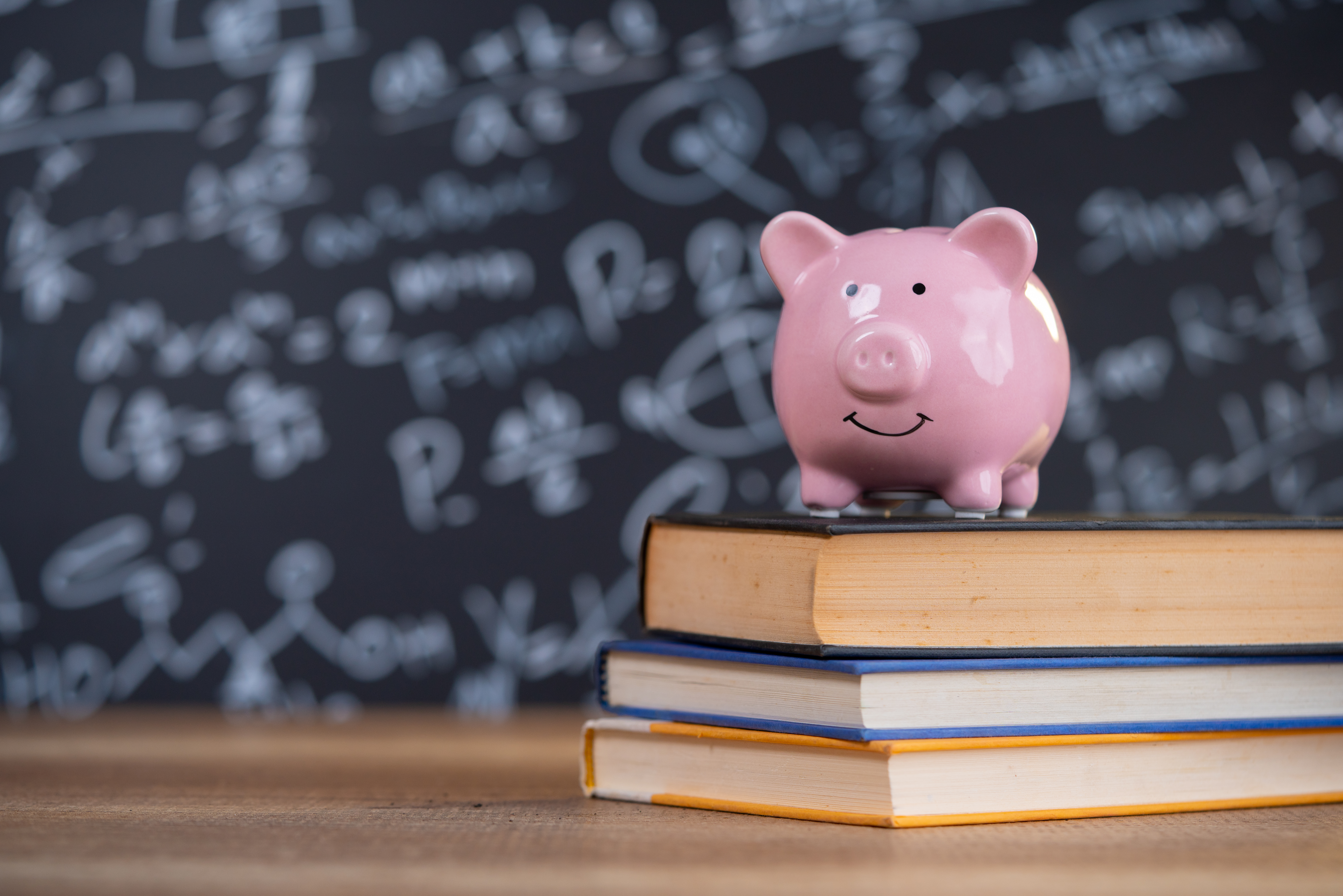 A piggy bank sits on top a stack of books in front of a chalkboard.