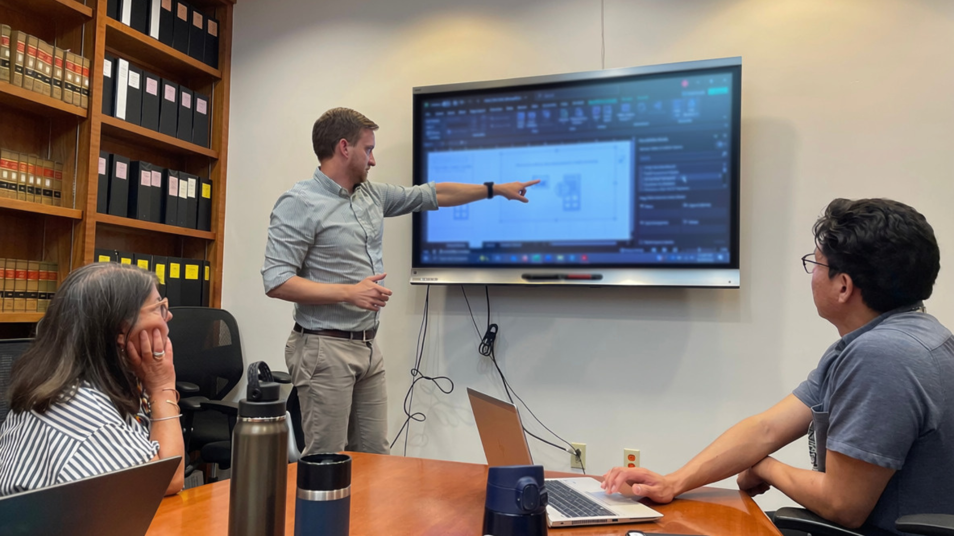 Two people sit at a table, looking at another person pointing at a screen.