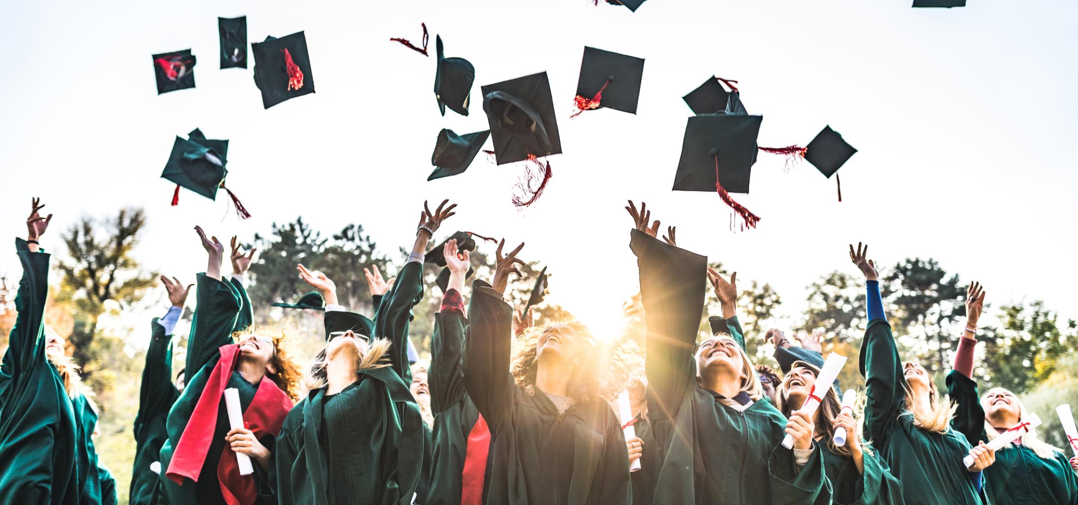 Students in graduation caps and gowns hold diplomas and smile as they throw their caps in the air.