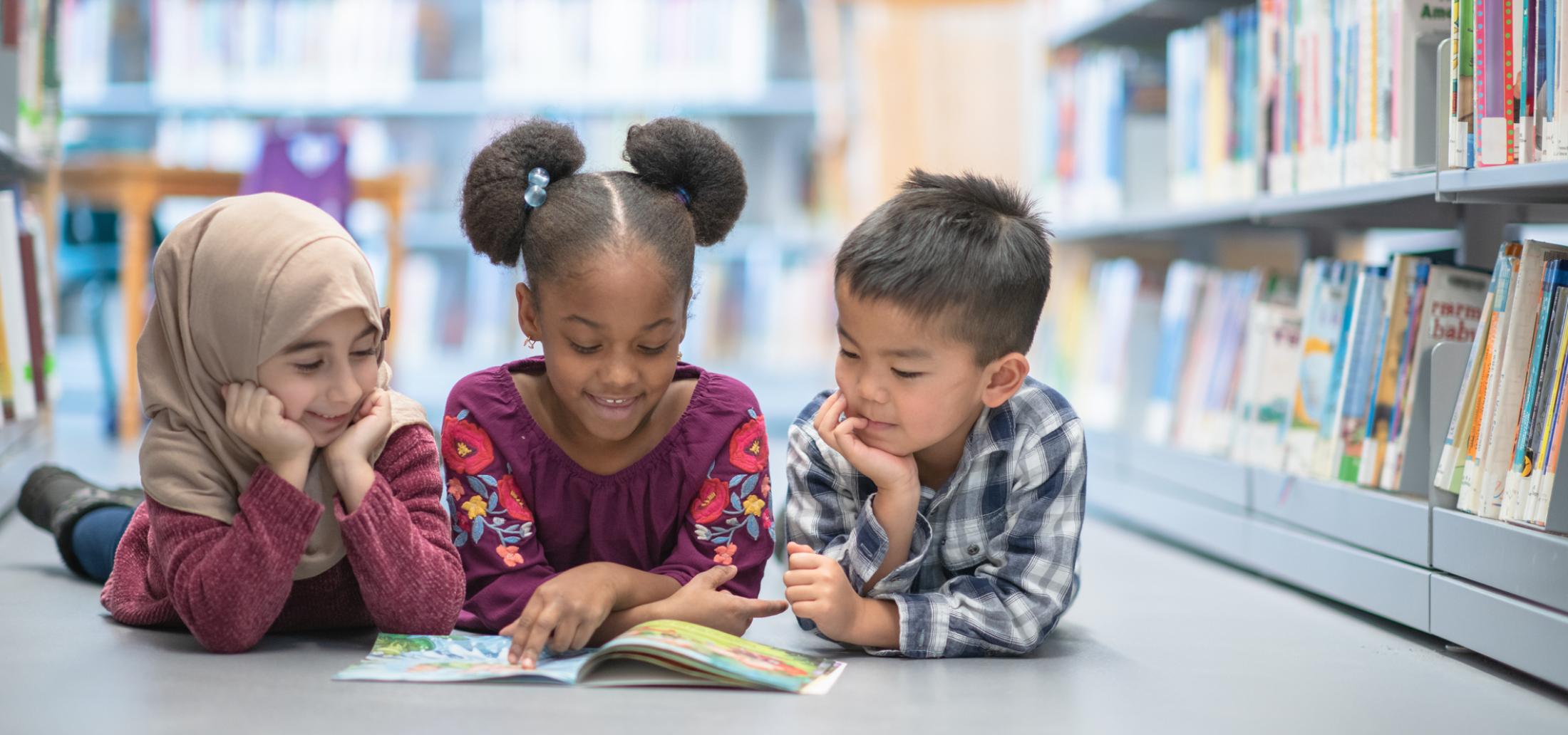 Three children laying on their stomachs between bookshelves, smiling and staring at a shared open book. 